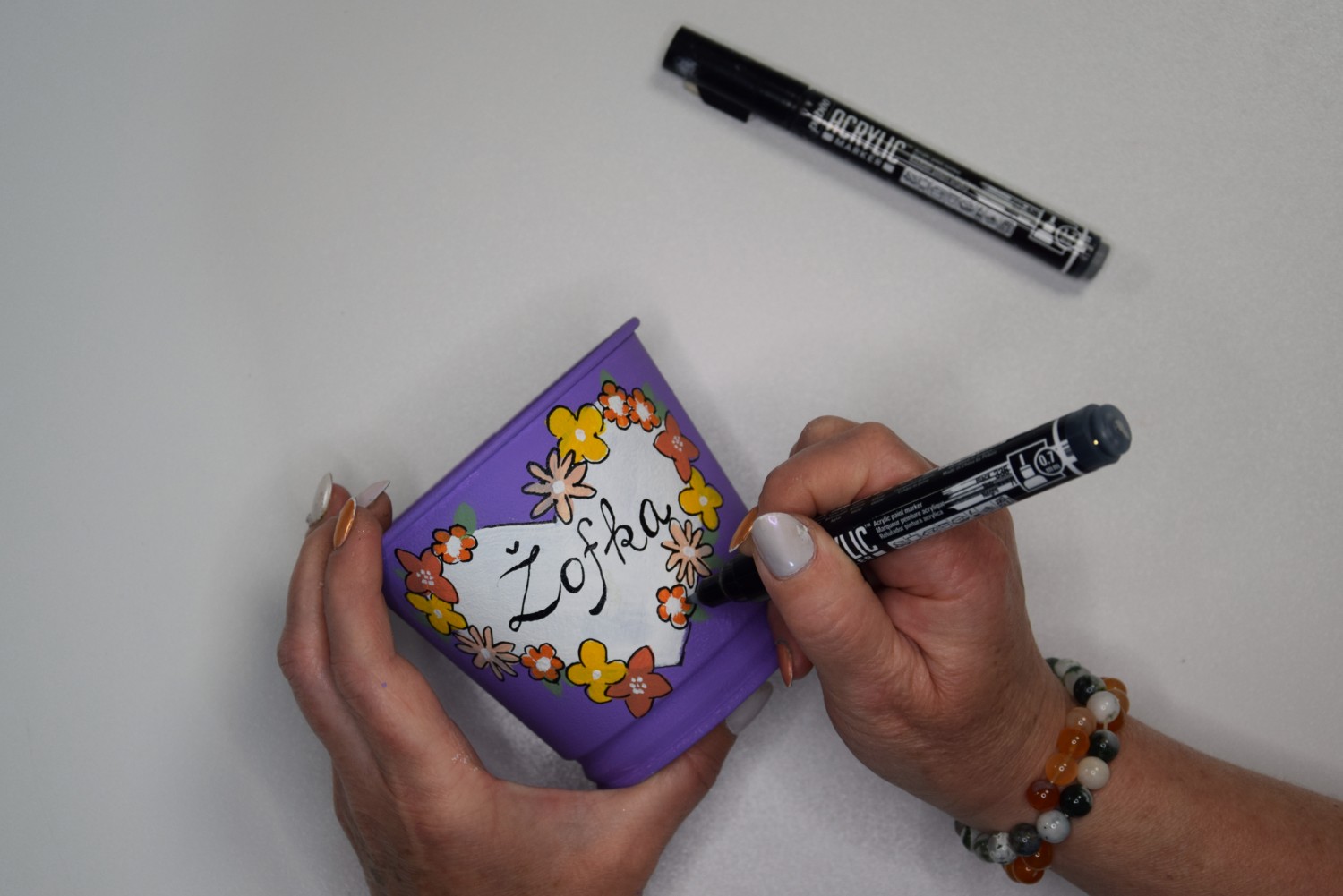 Žena pomocou čiernej akrylovej fixky maľuje na kvietkoch detaily a napíše aj slovo Žofka.